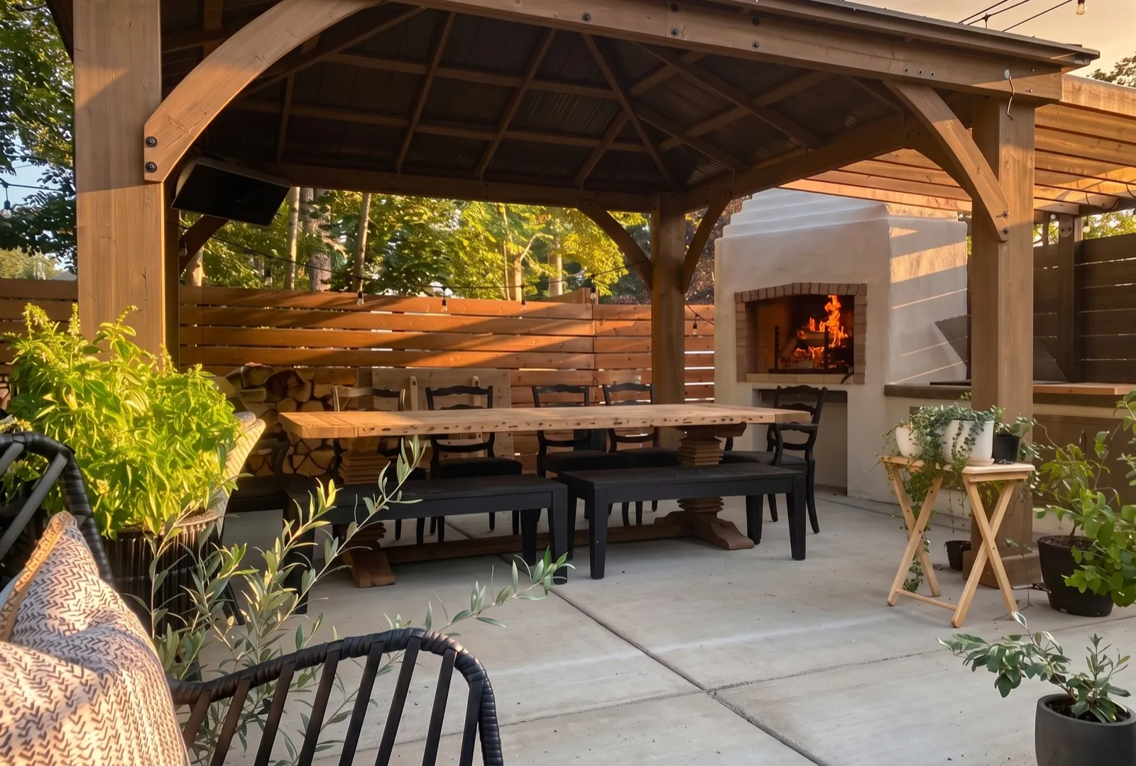 Timber pergola pavilion with a long reclaimed-wood dining table and a lit wood-fired fireplace at golden hour