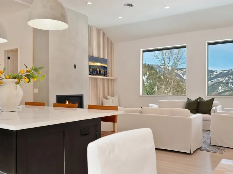 Remodeled living room with white slip-covered sofas, a fireplace, and floor-to-ceiling windows onto snow-capped mountains