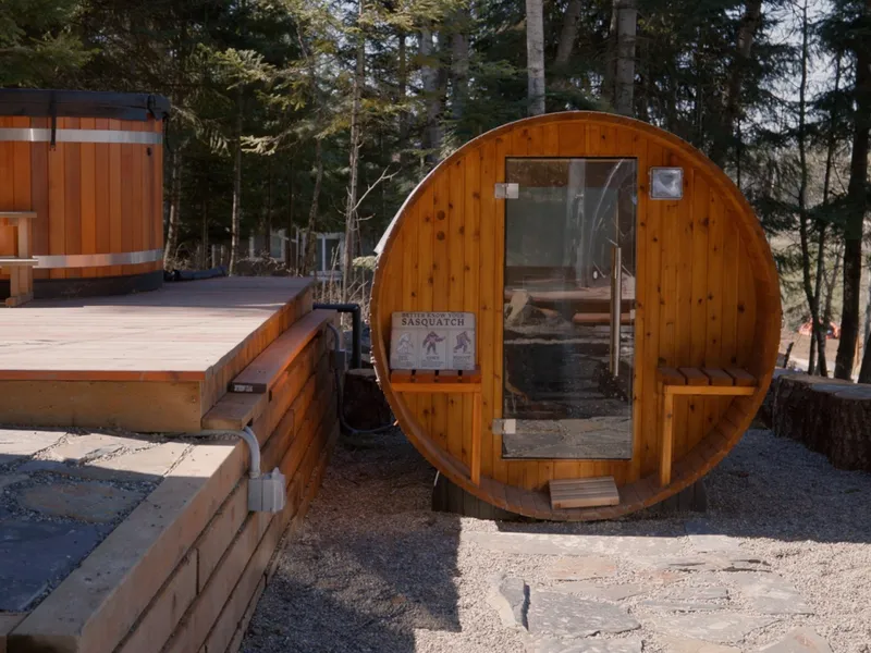 Cedar barrel sauna and round cold-plunge tub on a timber deck among pines in summer
